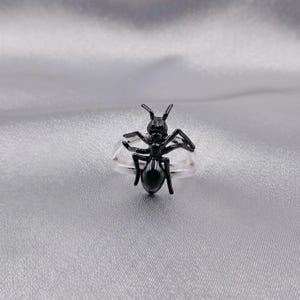 Può includere: Un anello nero a forma di formica. L'anello presenta un design dettagliato di formica tridimensionale con antenne, zampe e un corpo segmentato. La fascia dell'anello è trasparente, permettendo alla formica di risaltare. L'anello è posto su uno sfondo argentato.