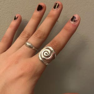 May include: Two silver rings are visible, one with a spiral design and the other with a teardrop shape. The hand also has dark nail polish.