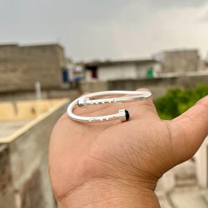 May include: A silver-colored bracelet with a nail-head design. The bracelet features a black gemstone at one end. The design includes small rectangular indentations along the nail head. The bracelet is shown on a hand.