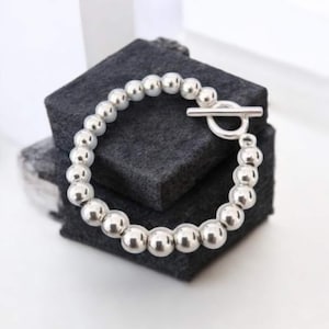 May include: A silver beaded bracelet with a toggle clasp. The bracelet features multiple round, shiny beads strung together. The clasp is a simple bar and loop design. The bracelet is displayed on a dark gray surface.