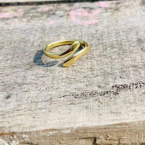Peut inclure: Une bague dorée au design ouvert unique. La bague présente un anneau lisse et incurvé qui s'enroule autour du doigt, avec une extrémité allongée légèrement plus épaisse. La bague est posée sur une surface en bois vieilli.