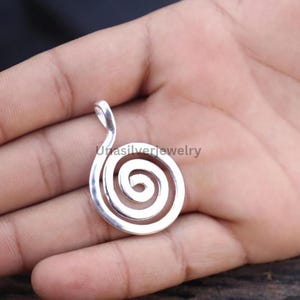 Puede incluir: Un colgante de metal plateado con un diseño en espiral, acabado liso y pulido, y una pequeña anilla integrada en la parte superior para su sujeción. El texto Unasilverjewelry es visible en el centro del colgante en espiral.