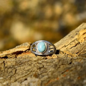 May include: A silver ring with an oval, light blue gemstone set in a gold bezel. The ring has an openwork design with decorative details. The ring is displayed on a piece of wood.
