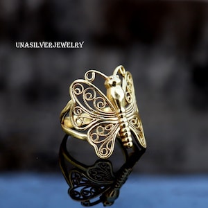 May include: A gold butterfly ring with detailed filigree patterns. The ring showcases a butterfly design with openwork wings and a textured body. The ring is presented on a reflective surface, with the text "UNASILVERJEWELRY" visible in the upper left corner.