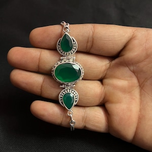 May include: A silver bracelet featuring three green gemstones. The bracelet has an oval-shaped center stone and two pear-shaped stones, all set in ornate silver settings. The bracelet is held against a dark background.