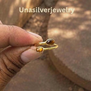 May include: A gold-toned open ring featuring two oval-shaped brown tiger's eye gemstones at each end of the band. The text Unasilverjewelry is visible at the top of the image. The ring has a polished finish and a bypass design.