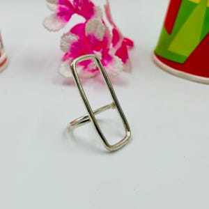 May include: A silver ring with a rectangular design. The ring features a vertical rectangle connected to a band. The ring is set against a white background with pink and white flowers and a colourful cup.