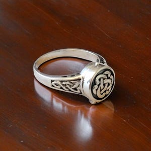 Könnte beinhalten: Ein silberner Ring mit einem keltischen Knotenmuster. Der Ring hat eine kreisförmige Oberfläche mit einem erhabenen Knotenmuster und aufwendigen Details am Band. Der Ring wird auf einer dunkelbraunen Oberfläche gezeigt.