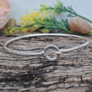 Op de afbeelding: Een zilverkleurige armband met een cirkelvormige sluiting. De armband wordt gepresenteerd op een rustiek houten oppervlak, met vage bloemen- en steenaccenten op de achtergrond. Een eenvoudig, elegant sieraad.