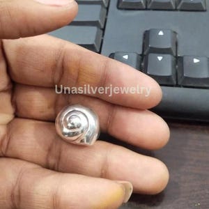 May include: A polished silver ring shaped like a spiraled seashell. The metal surface is smooth and reflective, featuring a classic nautilus-inspired coil design. The text Unasilverjewelry is visible in the background near the silver ring.