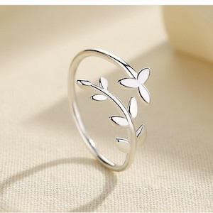 May include: A silver-colored ring with a floral design. The ring features a delicate vine with leaves and a flower. The ring is made of a shiny metal and is set against a neutral background. The ring is a piece of jewelry.