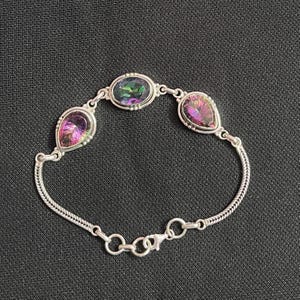 May include: A silver chain bracelet featuring three iridescent, multi-colored gemstones. The center stone is oval-shaped, while the two flanking stones are pear-shaped. The bracelet includes a delicate chain link design and a secure lobster clasp.