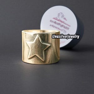May include: A gold-coloured ring featuring a raised star design. The ring is cylindrical with a textured surface, and the star is centred on the band. It's set against a dark background with a partially visible round container.