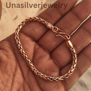 Puede incluir: Una pulsera de cadena trenzada de color cobre con un acabado metálico y un cierre de gancho. El texto Unasilverjewelry es visible en la esquina superior izquierda de la imagen. La pulsera tiene un patrón repetitivo de eslabones trenzados.