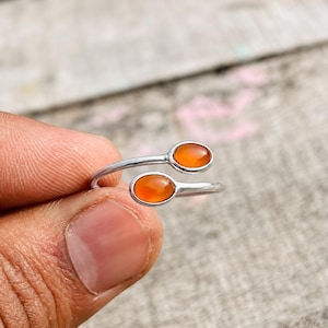 May include: A bypass ring crafted from silver-toned metal featuring two oval-shaped, smooth orange cabochon stones set at each end of the open band. The design showcases a sleek, polished finish with the two stones positioned to overlap slightly.