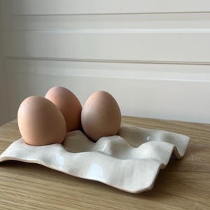 May include: Three brown eggs rest in a cream-colored ceramic egg holder. The holder has a wavy design with four depressions for the eggs. The eggs are a light brown color and are placed on a wooden surface.