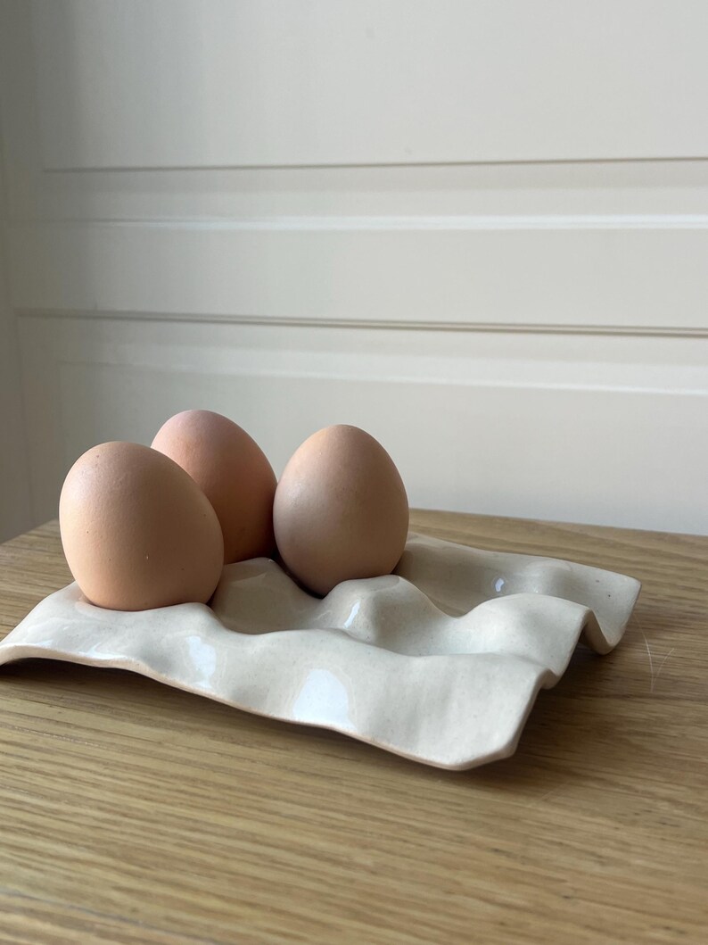 May include: Three brown eggs rest in a cream-colored ceramic egg holder. The holder has a wavy design with four depressions for the eggs. The eggs are a light brown color and are placed on a wooden surface.