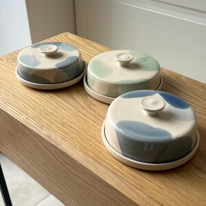May include: Three ceramic butter dishes with lids. Each dish has a round base and a domed lid with a small knob. The lids are decorated with blue, green, and gray abstract patterns on a cream background. The dishes are displayed on a wooden surface.