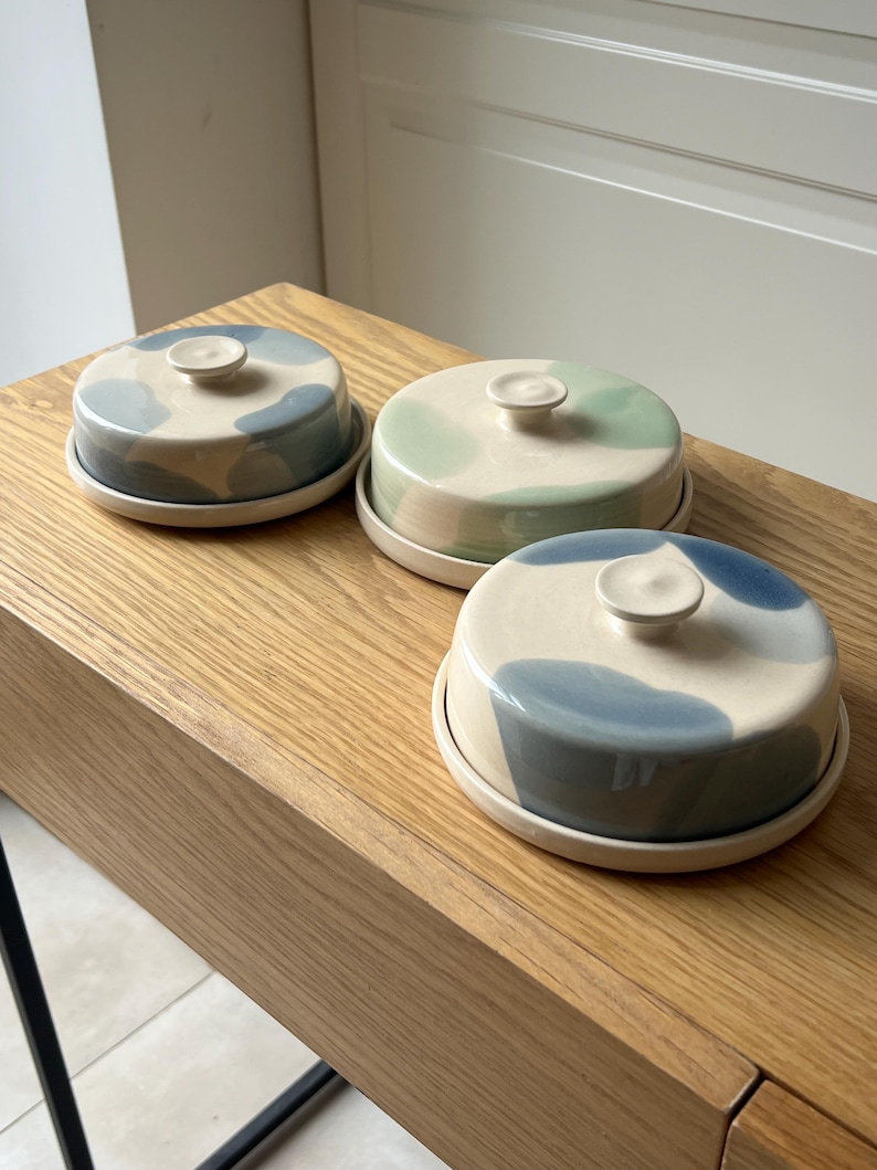 May include: Three ceramic butter dishes with lids. Each dish has a round base and a domed lid with a small knob. The lids are decorated with blue, green, and gray abstract patterns on a cream background. The dishes are displayed on a wooden surface.