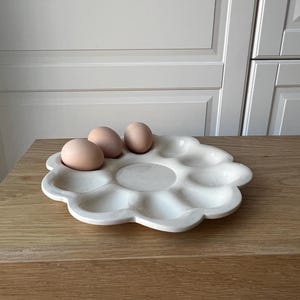 May include: A white ceramic egg holder in a flower shape, holding three brown eggs. The egg holder has multiple compartments and sits on a wooden surface. The background features a neutral-colored wall and cabinet.