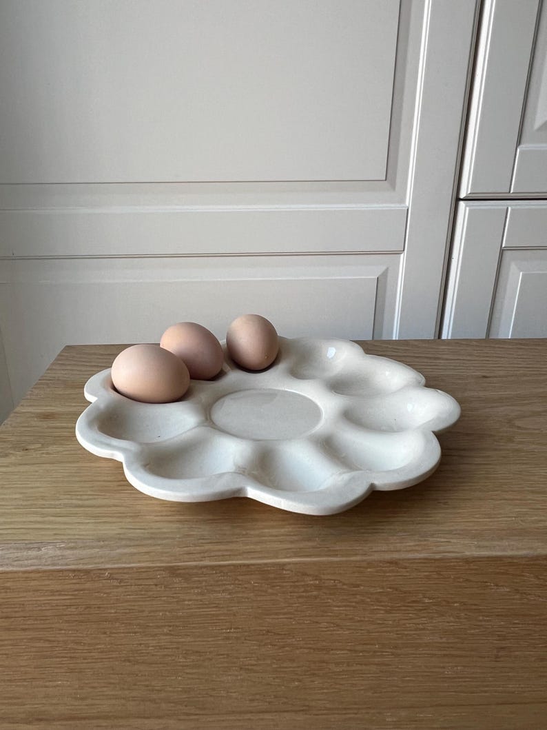 May include: A white ceramic egg holder in a flower shape, holding three brown eggs. The egg holder has multiple compartments and sits on a wooden surface. The background features a neutral-colored wall and cabinet.