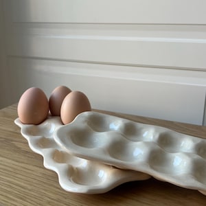 May include: Two off-white ceramic egg trays, each with six egg-shaped indentations, are stacked on a wooden surface. Three brown eggs rest in the top tray. The trays have a wavy, organic design.