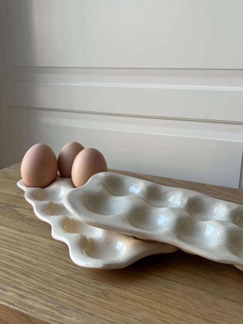 May include: Two off-white ceramic egg trays, each with six egg-shaped indentations, are stacked on a wooden surface. Three brown eggs rest in the top tray. The trays have a wavy, organic design.