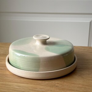 May include: A ceramic butter dish with a lid. The dish is round with a cream-colored base and lid. The lid features a light green watercolor design. The knob on top is cream-colored. The dish sits on a wooden surface.