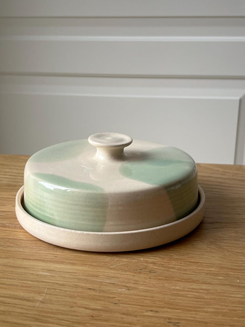 May include: A ceramic butter dish with a lid. The dish is round with a cream-colored base and lid. The lid features a light green watercolor design. The knob on top is cream-colored. The dish sits on a wooden surface.