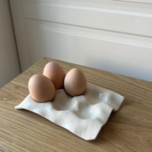 May include: A cream-colored ceramic egg holder with three brown eggs. The holder has a wavy design with four indentations. The eggs are light brown and sit on a wooden surface.