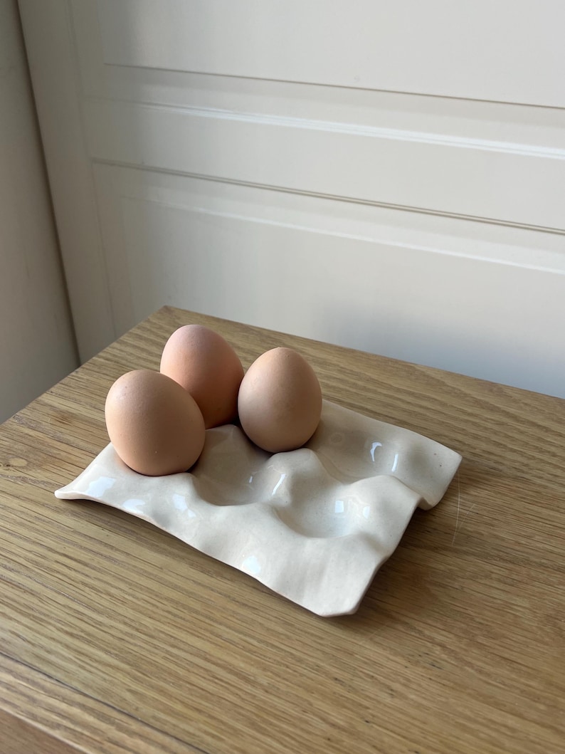 May include: A cream-colored ceramic egg holder with three brown eggs. The holder has a wavy design with four indentations. The eggs are light brown and sit on a wooden surface.