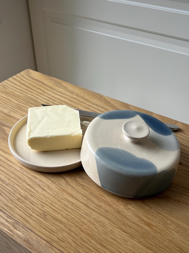 May include: A ceramic butter dish with a lid, featuring blue abstract designs, sits next to a small plate holding a square of butter. A butter knife rests on the plate. The items are on a wooden surface.