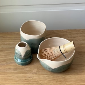 Pode incluir: Um conjunto de chá matcha de cerâmica em branco e verde-azulado. O conjunto inclui uma tigela com um batedor de bambu, um vaso pequeno e uma chávena alta. As peças estão dispostas sobre uma superfície de madeira.