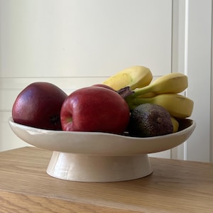 Könnte beinhalten: Eine cremefarbene Obstschale aus Keramik, gefüllt mit roten Äpfeln, Bananen und einer Avocado. Die Schale steht auf einer Holzoberfläche, mit einer weißen Tür im Hintergrund. Die Obstschale ist ein dekorativer Artikel für die Küche.