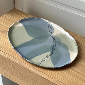 Pode incluir: Um prato oval de cerâmica com uma borda irregular e ondulada. A superfície apresenta formas abstratas sobrepostas em tons de verde sálvia e azul acinzentado, acentuadas por uma linha preta fina desenhada à mão que atravessa o centro.