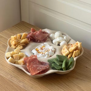 Könnte beinhalten: Ein beigefarbenes Serviertablett mit mehreren Fächern, gefüllt mit verschiedenen Lebensmitteln. Enthält Käsewürfel, Aufschnitt, Mozzarellabällchen sowie grüne und orangefarbene Lebensmittel. Das Tablett steht auf einer Holzoberfläche.
