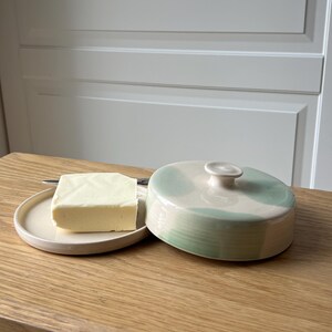 May include: A ceramic butter dish with a lid, featuring a light green and white design. A square of butter sits on a small, round plate next to the dish. A small knife is also on the plate. The butter dish is on a wooden surface.