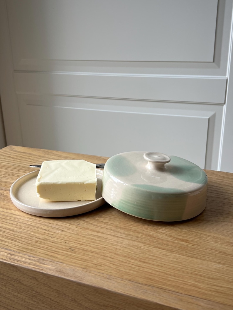 May include: A ceramic butter dish with a lid, featuring a light green and white design. A square of butter sits on a small, round plate next to the dish. A small knife is also on the plate. The butter dish is on a wooden surface.