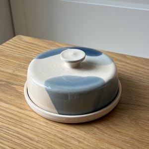 May include: A ceramic butter dish with a lid, featuring a cream base and blue abstract designs. The dish has a round shape with a small knob on top of the lid. The butter dish sits on a matching round base.