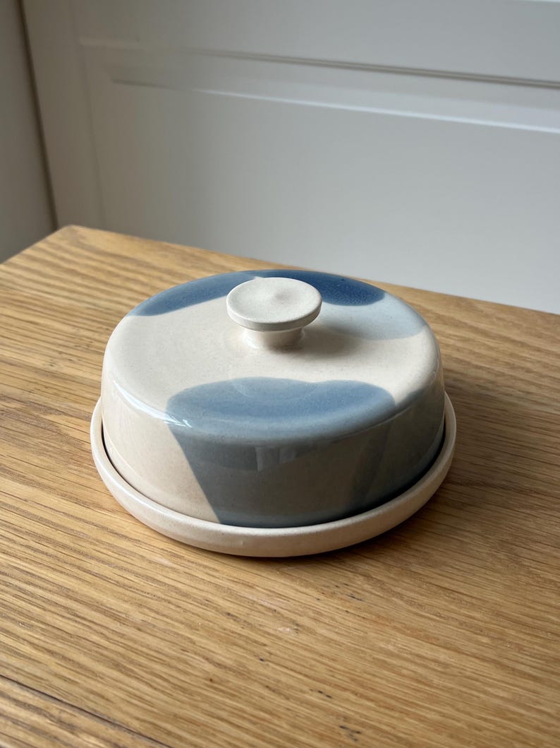 May include: A ceramic butter dish with a lid, featuring a cream base and blue abstract designs. The dish has a round shape with a small knob on top of the lid. The butter dish sits on a matching round base.
