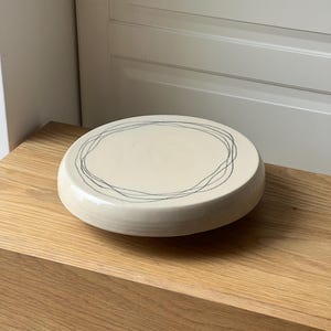May include: A circular, cream-coloured ceramic dish featuring a hand-drawn, black, circular design. The dish sits on a light-coloured wooden surface. The design is a simple, yet elegant, decorative element.