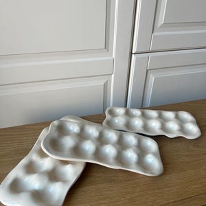 May include: Three off-white ceramic egg trays on a wooden surface. Each tray has rounded indentations to hold eggs. The trays are rectangular with a slightly irregular shape, and the background features a white cabinet.