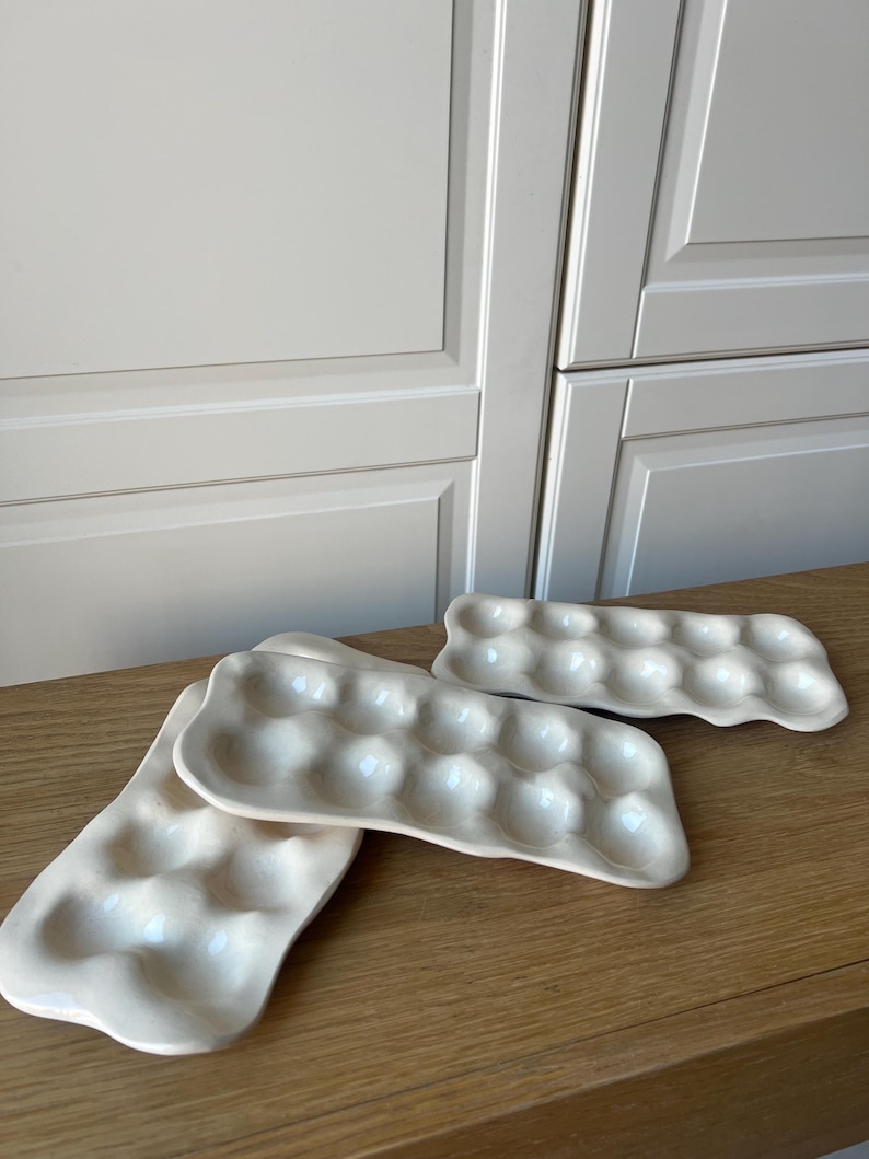 May include: Three off-white ceramic egg trays on a wooden surface. Each tray has rounded indentations to hold eggs. The trays are rectangular with a slightly irregular shape, and the background features a white cabinet.