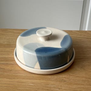 May include: A ceramic butter dish with a lid and base. The lid is decorated with blue and off-white abstract shapes. The dish has a small, round knob on top for easy lifting. The base is a simple, round plate.