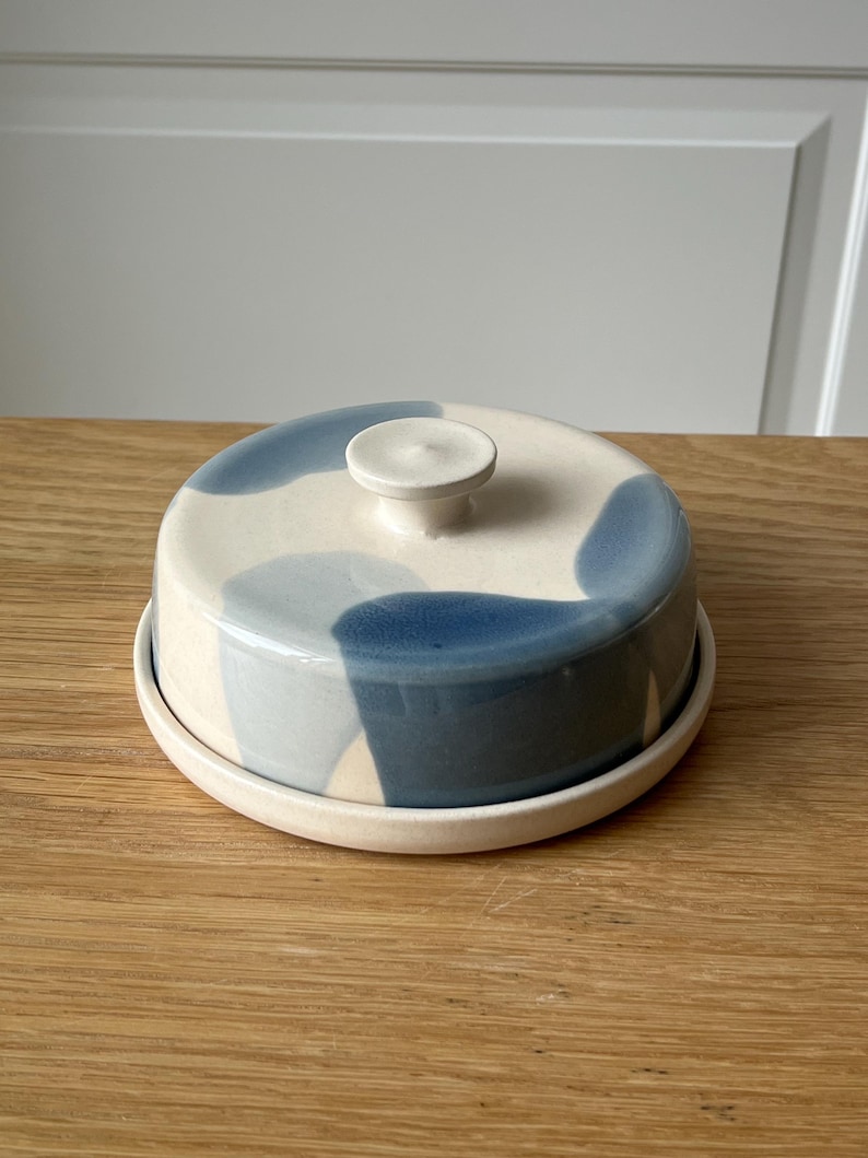 May include: A ceramic butter dish with a lid and base. The lid is decorated with blue and off-white abstract shapes. The dish has a small, round knob on top for easy lifting. The base is a simple, round plate.