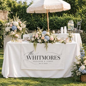 May include: A white table runner featuring the black printed text The WHITMORES, ELEANOR & JAMES, and 09.12.2026. The runner is draped over a white tablecloth on a table decorated with a floral centerpiece of blue, pink, and white flowers.