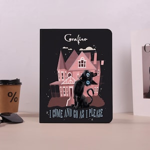 May include: Black notebook with a cartoon illustration of a black cat with blue eyes in front of a pink house. The text "Coraline" is above the house, and "I COME AND GO AS I PLEASE" is below.