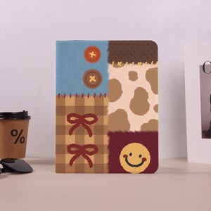 Pode incluir: Uma capa retangular para tablet com design de patchwork. O lado esquerdo exibe uma seção azul com dois botões laranja e uma seção xadrez marrom com dois laços vermelho-escuros. O lado direito mostra um padrão de manchas marrons e um rosto sorridente amarelo.