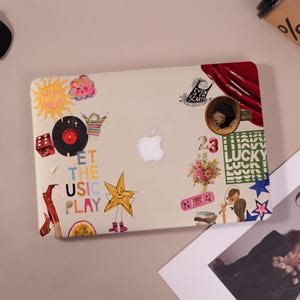 Puede incluir: Un portátil beige con pegatinas coloridas, incluyendo un sol, un disco, una estrella y la frase "LET THE MUSIC PLAY". Una taza de café está en la esquina superior derecha. El logotipo de Apple es visible.