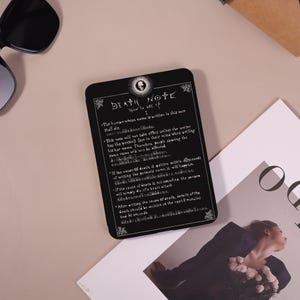 May include: Black rectangular Death Note card with white text and decorative borders. The card features the title "DEATH NOTE" and instructions on how to use it. A skull illustration is at the top. The card is on a light-colored surface.